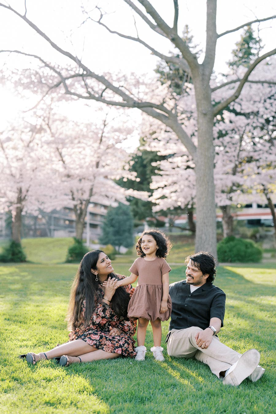 A family enjoying a sunny day in a park surrounded by blooming cherry blossom trees, with a mother sitting on the grass, a child standing, and a father nearby, all smiling and embracing the peaceful outdoor setting.