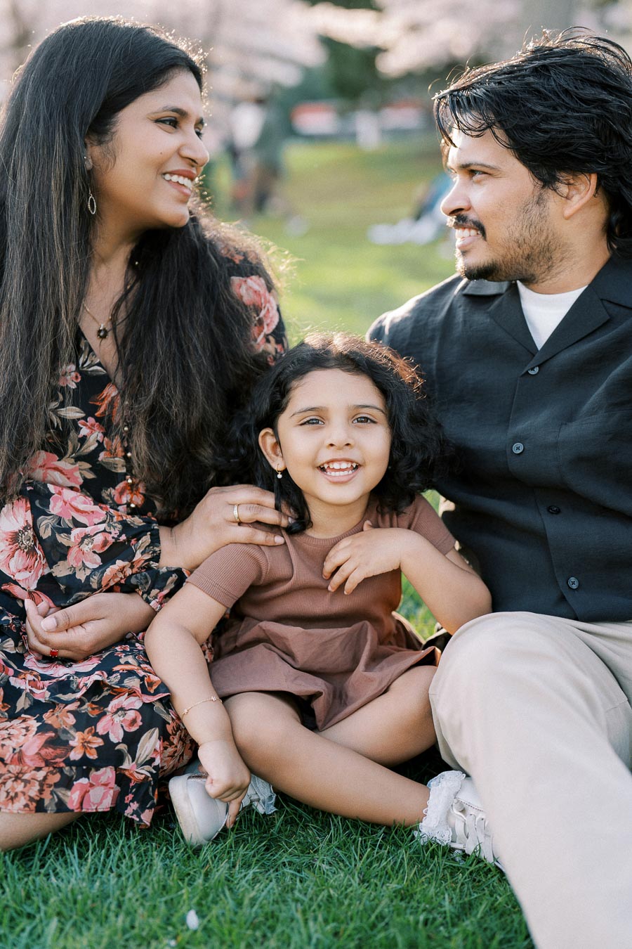 A happy family enjoying a day outdoors, sitting on green grass. The mother, wearing a floral dress, and the father, in a black shirt, smile warmly at their young daughter, who is smiling joyfully between them. Bright and sunny day, highlighting family bonding and joy.