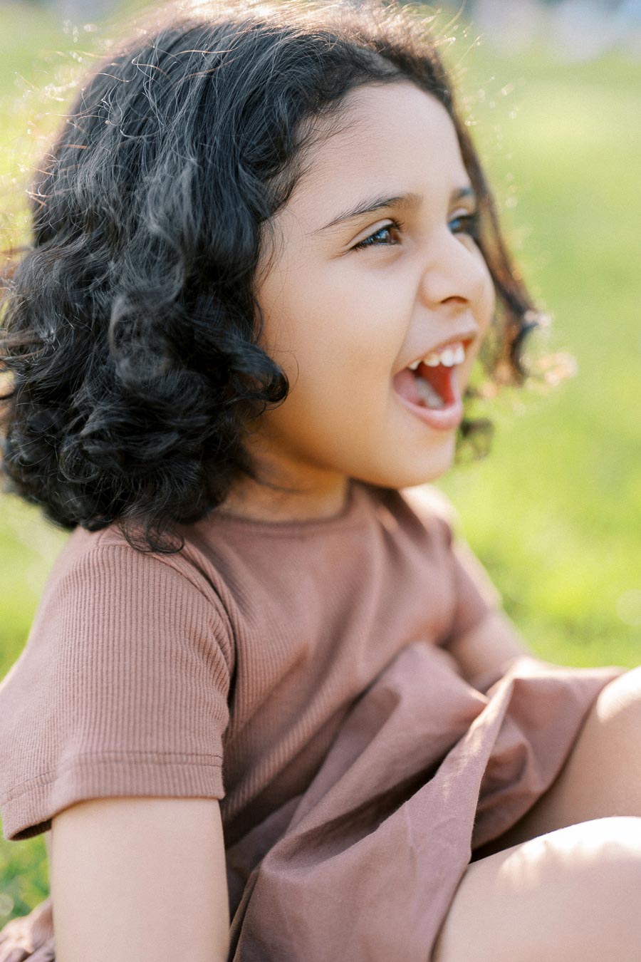 A joyful child with curly hair sitting outdoors and smiling brightly on a sunny day, enjoying the vibrant green surroundings.