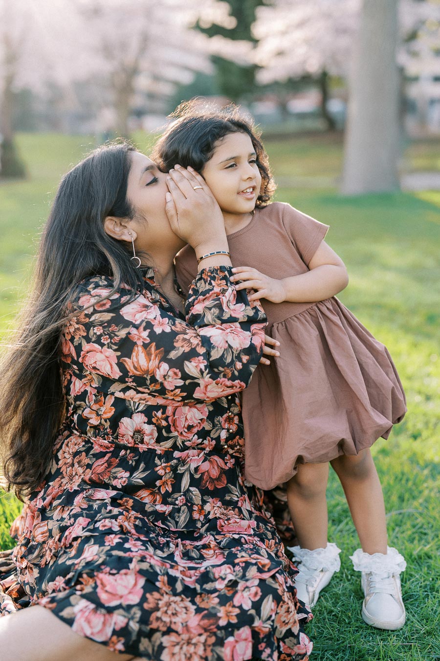 A woman in a floral dress whispers to a young girl in a brown dress as they sit on the grass in a sunlit park, surrounded by blooming trees.