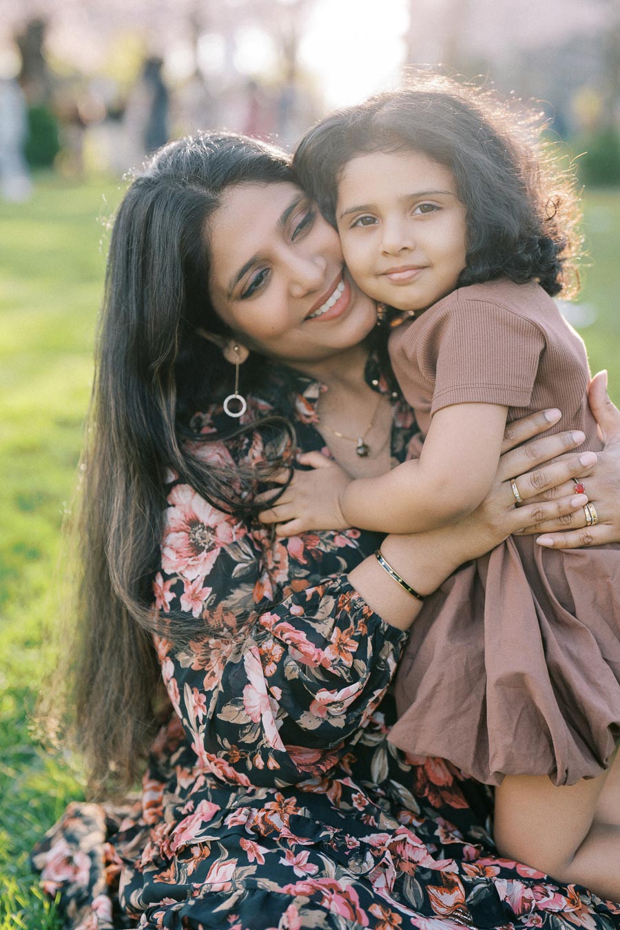 A mother lovingly embraces her daughter on a sunny day in a park, both smiling warmly. The mother wears a floral dress, and the child wears a brown outfit. The natural backdrop features greenery and soft sunlight, creating a joyful and serene atmosphere. Perfect depiction of family bonding and happiness.