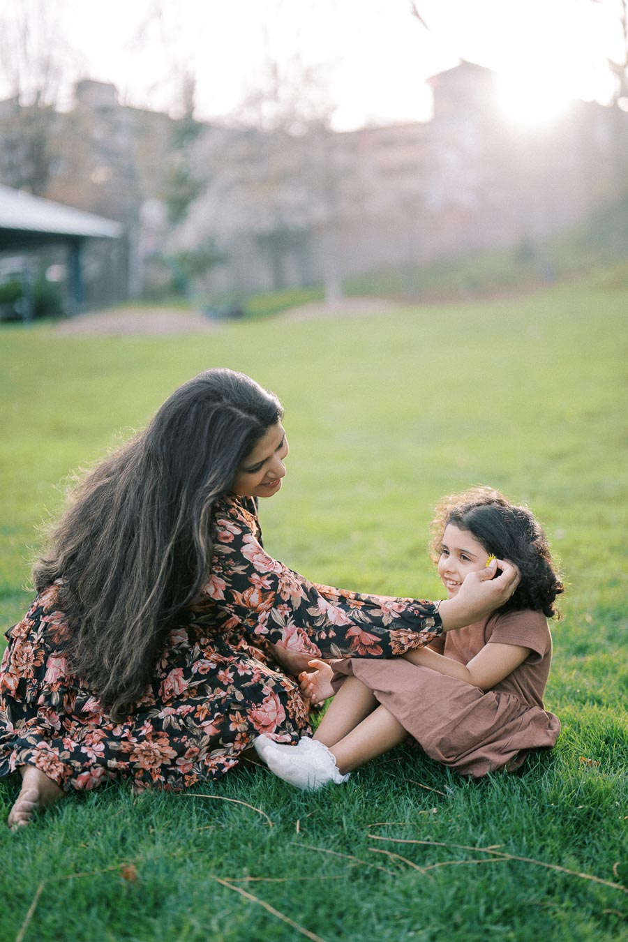 Woman and young girl sitting on green grass in a park, wearing floral and brown dresses, sharing a joyful moment, with sunlight filtering through trees in the background.