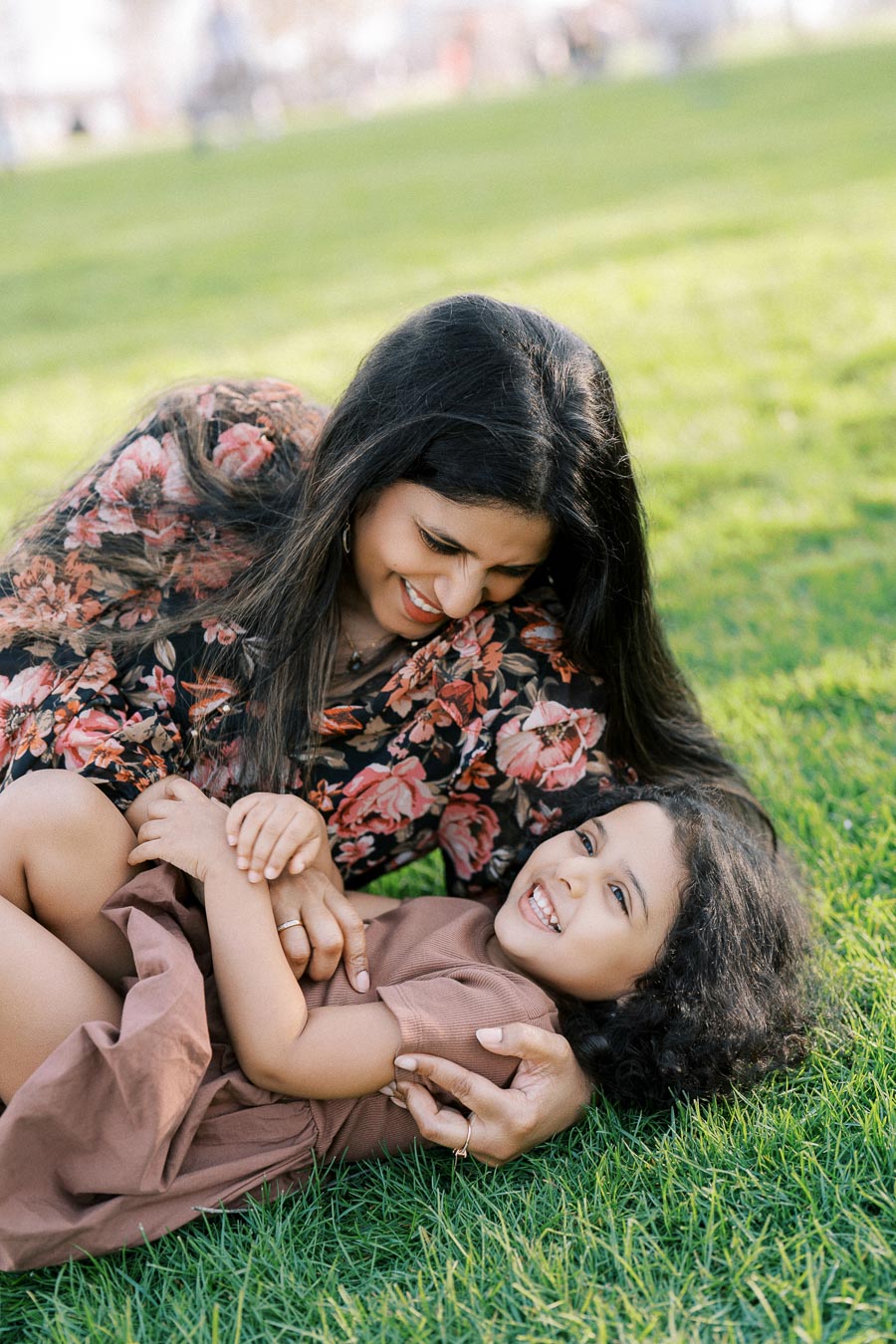 A woman in a floral dress smiling and playing with a laughing child on green grass during a sunny day in a park.