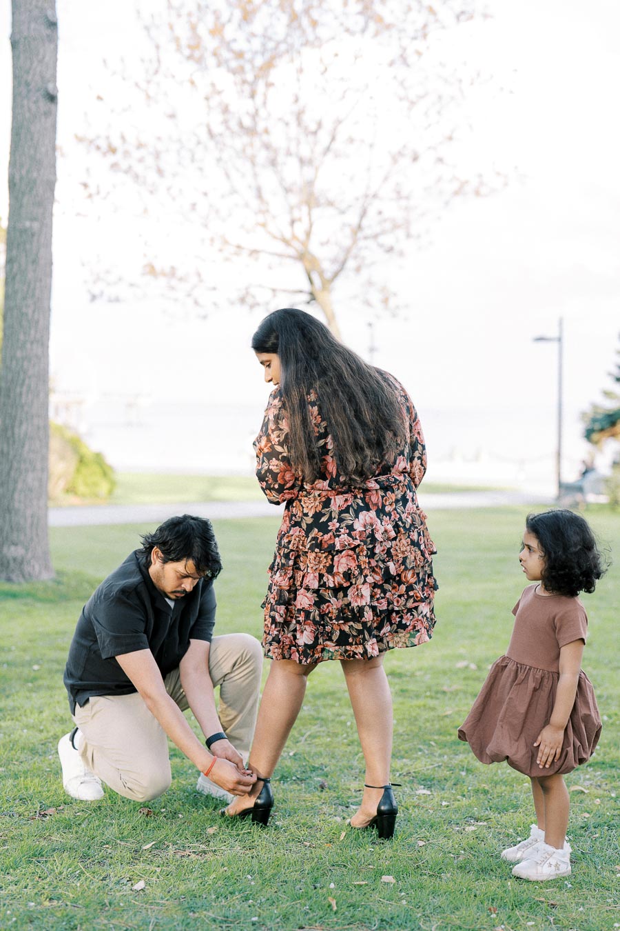 A man kneeling on the grass helps a woman in a floral dress with her shoe, while a young girl in a brown dress watches. The scene is set in a park with trees and a clear sky.