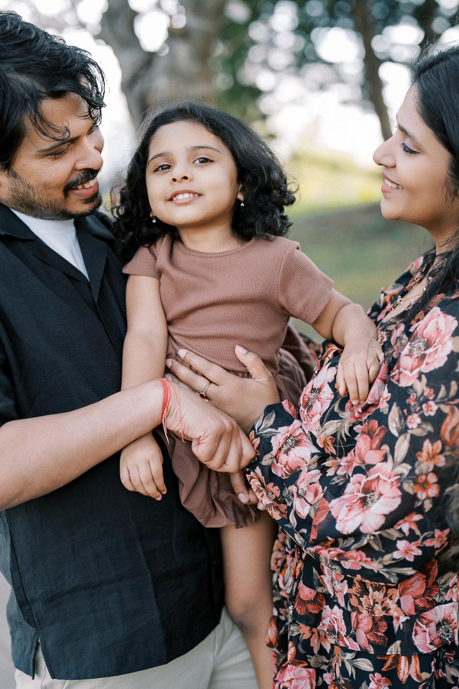 A happy family outdoors, featuring a mother and father holding their young daughter, surrounded by greenery and trees in a park setting.