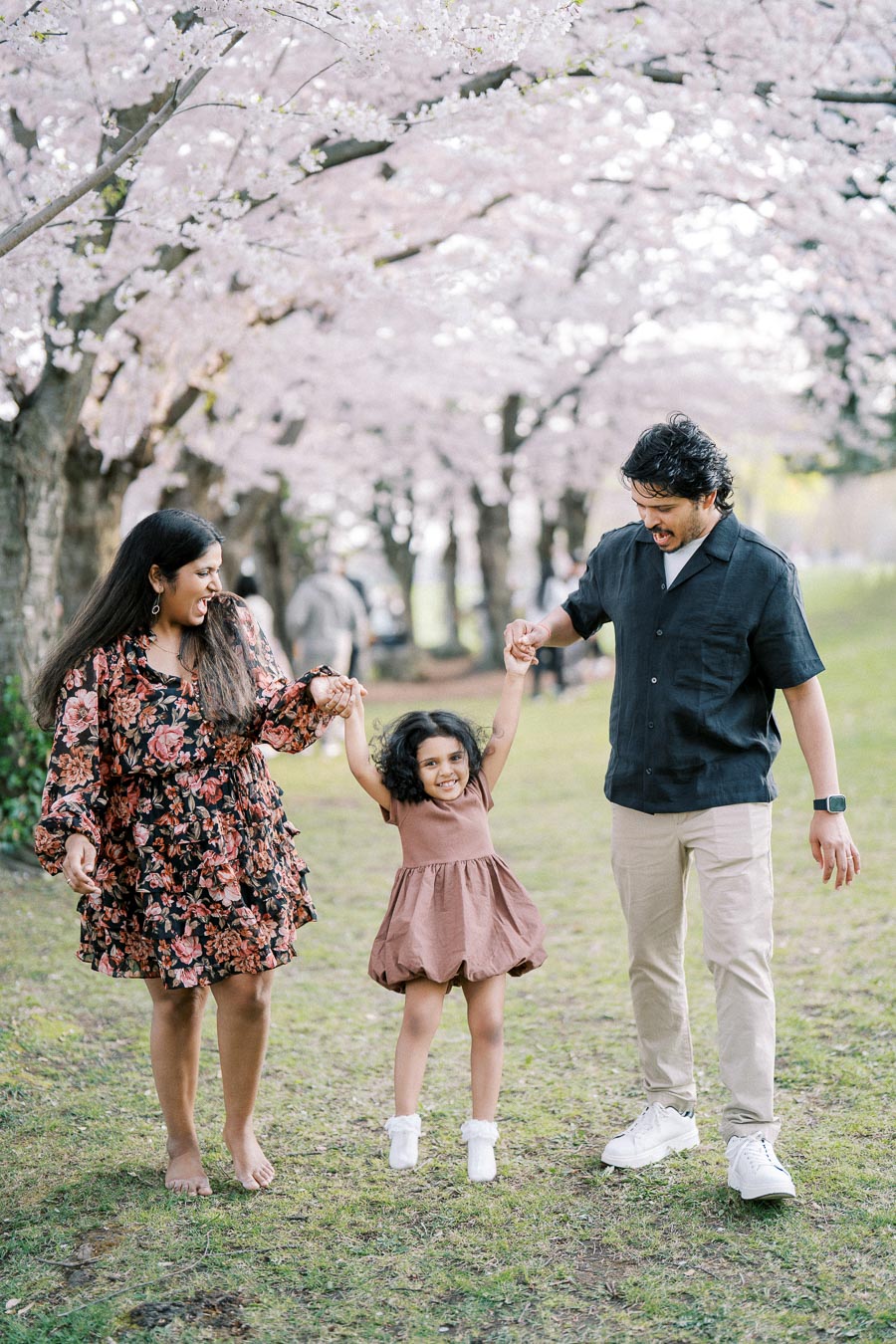 A joyful family of three enjoying a spring day under blooming cherry blossom trees, with parents holding their daughter's hands as she jumps in the middle, smiling and surrounded by soft pink petals.