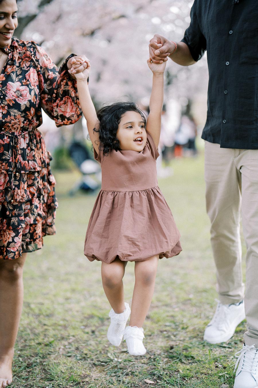 A joyful young girl in a brown dress is swinging between two adults holding her hands, surrounded by blooming cherry blossoms in a park.