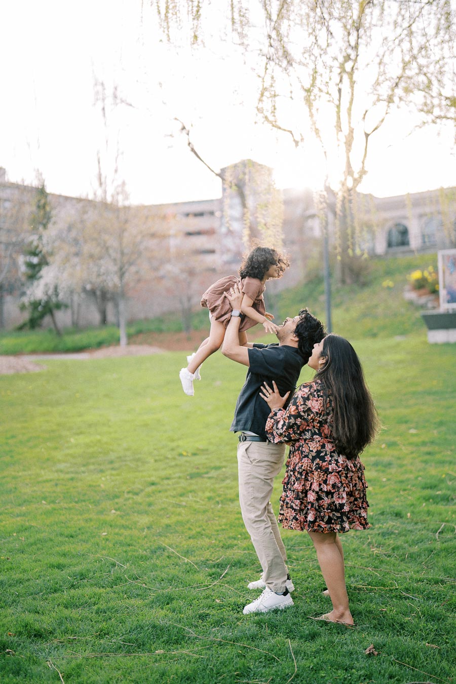 A happy family enjoying a sunny day at the park, with a father playfully lifting his child in the air and the mother lovingly watching beside them on lush green grass, surrounded by trees and a building in the background.