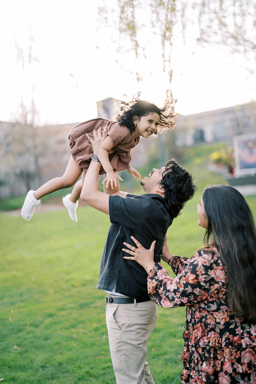A joyful family moment in a park with a father playfully lifting his smiling daughter into the air, while the mother happily looks on, all enjoying a sunny day outdoors.