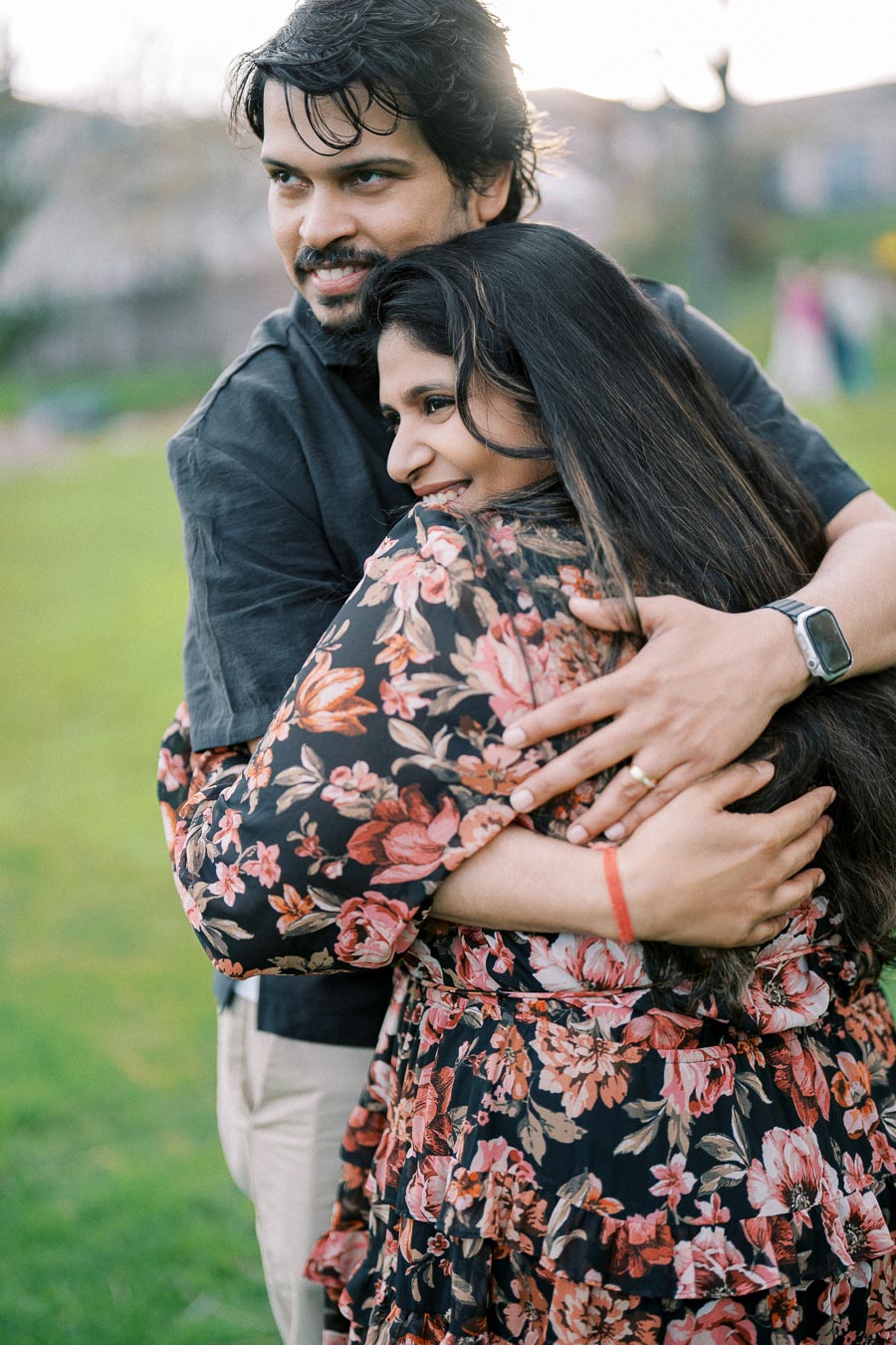 A couple embracing and smiling outdoors on a sunny day, with a background of green grass and blurred scenery. The woman is wearing a floral dress and the man is in a dark shirt, showcasing a joyful and romantic moment in a natural setting.