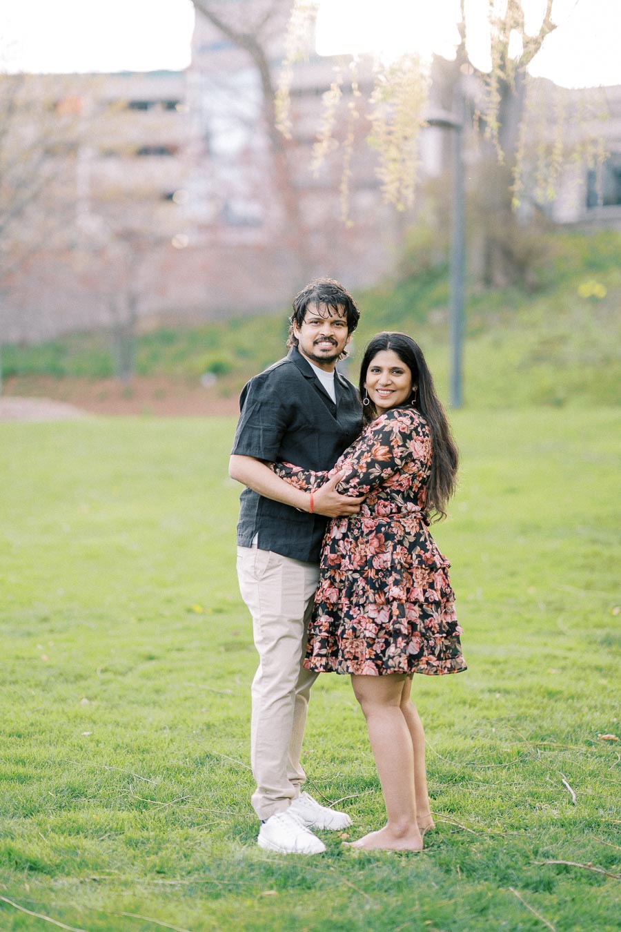 A couple embracing in a scenic park with lush green grass and soft sunlight filtering through trees, conveying a warm and romantic atmosphere.