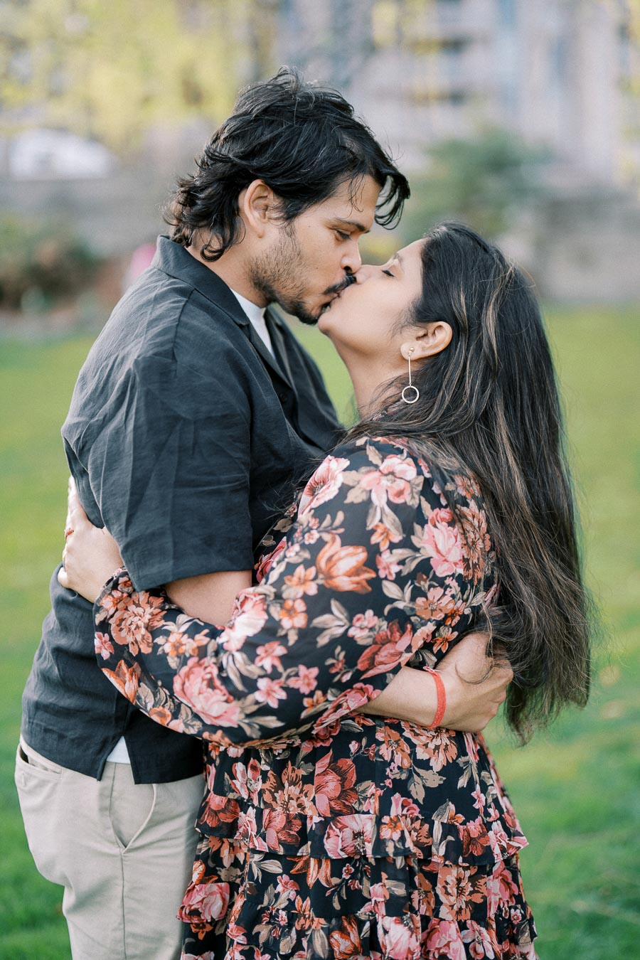 A couple shares a romantic kiss in a park setting, with the man wearing a dark shirt and the woman in a floral dress, capturing a tender moment outdoors.