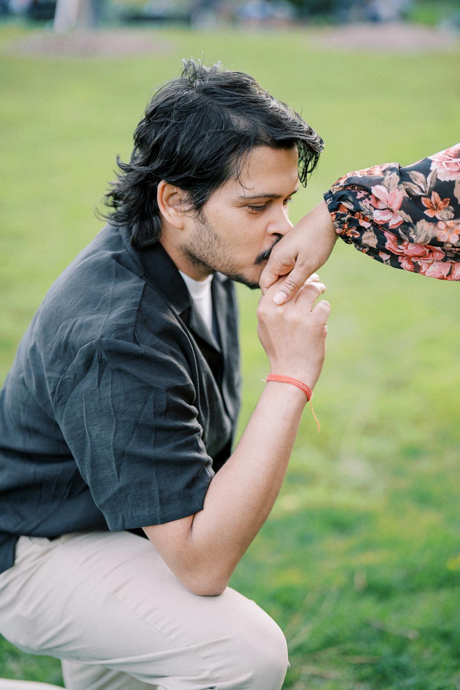 A man gently kisses a woman's hand while kneeling in a green outdoor setting, creating a romantic and affectionate moment.