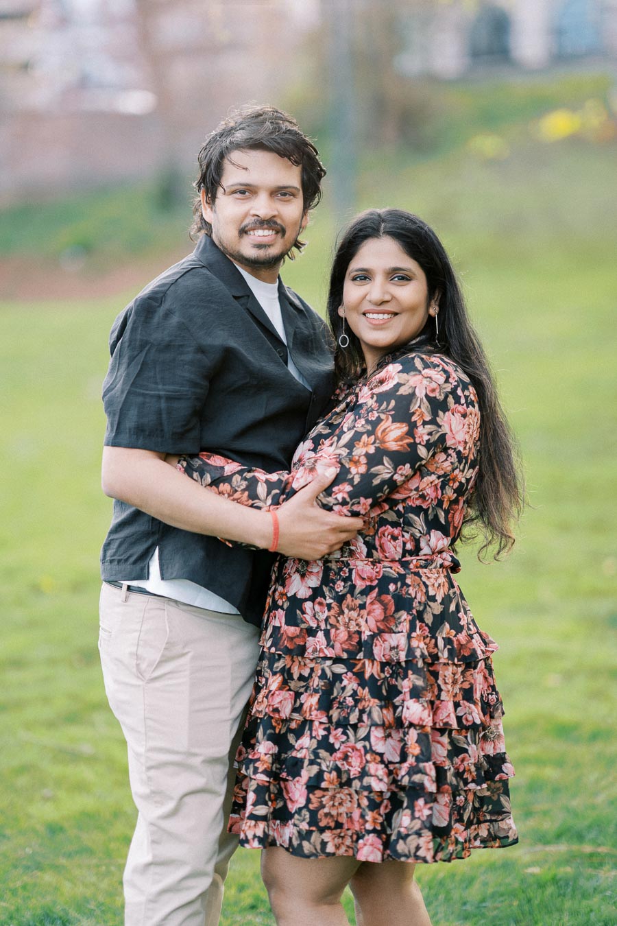 A smiling couple embraces in a sunny park, with the man wearing a black shirt and the woman in a floral dress, standing on lush green grass.