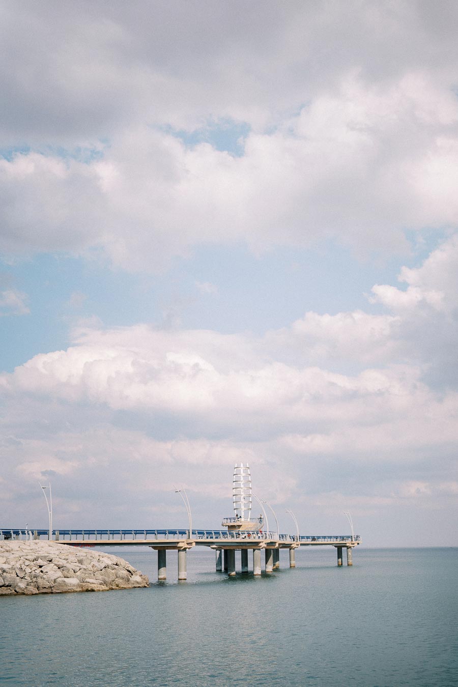 Scenic view of a modern pier extending into the calm ocean, under a partly cloudy sky, with urban architecture and lampposts lining the structure.