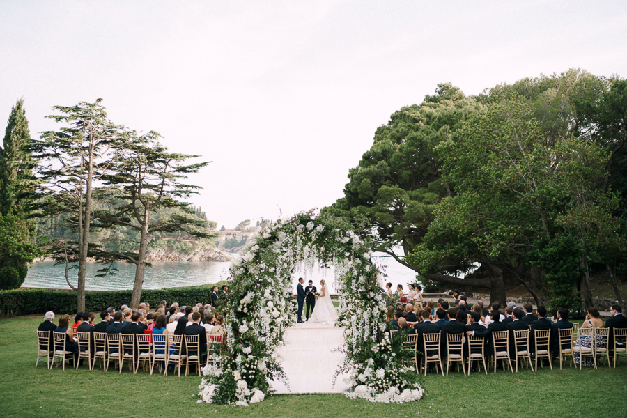 Outdoor wedding ceremony by a scenic lake, featuring a floral archway, bride and groom standing at the altar, and guests seated on both sides surrounded by lush greenery.
