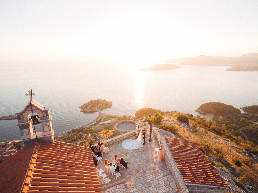Aerial view of a scenic outdoor wedding ceremony on a cliffside overlooking the ocean at sunset, with guests seated and a couple standing under a floral arch, surrounded by breathtaking coastal landscape and islands.