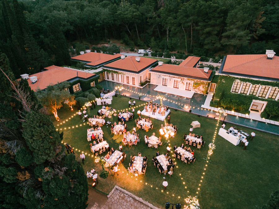 Aerial view of an elegant outdoor wedding reception with round tables and string lights, set on the lawn of a luxurious estate surrounded by greenery and modern architecture.