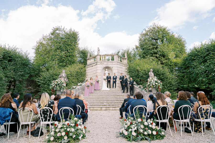 Upscale outdoor wedding ceremony at historic venue with classic stone architecture and lush greenery, featuring bride, groom, and wedding party on steps, surrounded by seated guests.