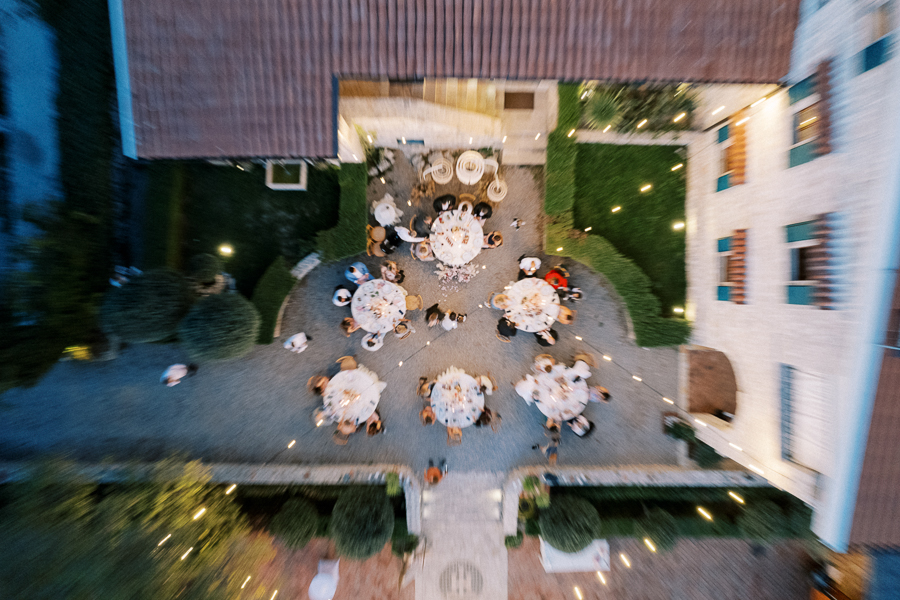 Aerial view of an elegant outdoor dinner event with multiple round tables surrounded by guests in a courtyard setting, accentuated by atmospheric string lights, and bordered by lush greenery and a charming building.