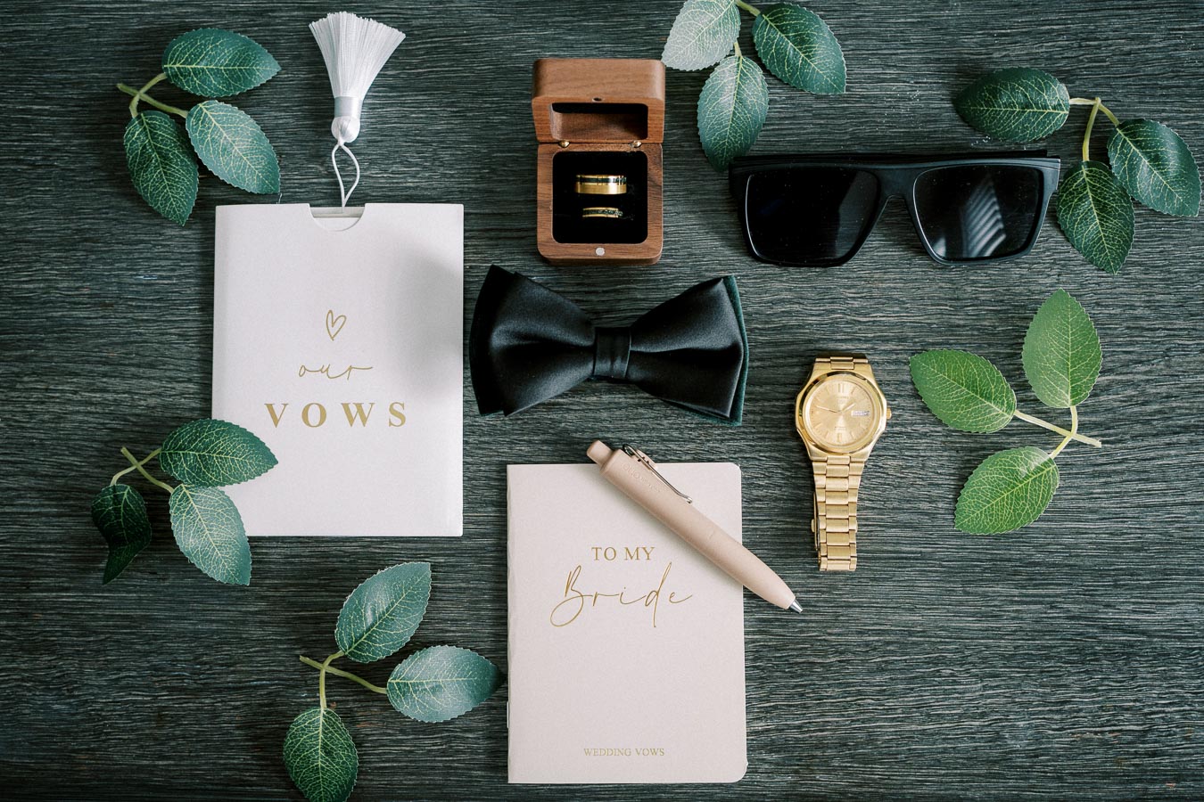Wedding accessories flat lay with vow books, black bow tie, gold watch, sunglasses, and wedding rings in a wooden box on a dark wooden background adorned with green leaves.