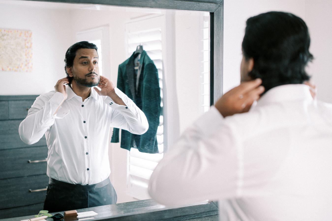 Man adjusts his collar while looking in the mirror, wearing a white dress shirt and preparing for a formal event, with a suit jacket visible in the background, in a well-lit room.