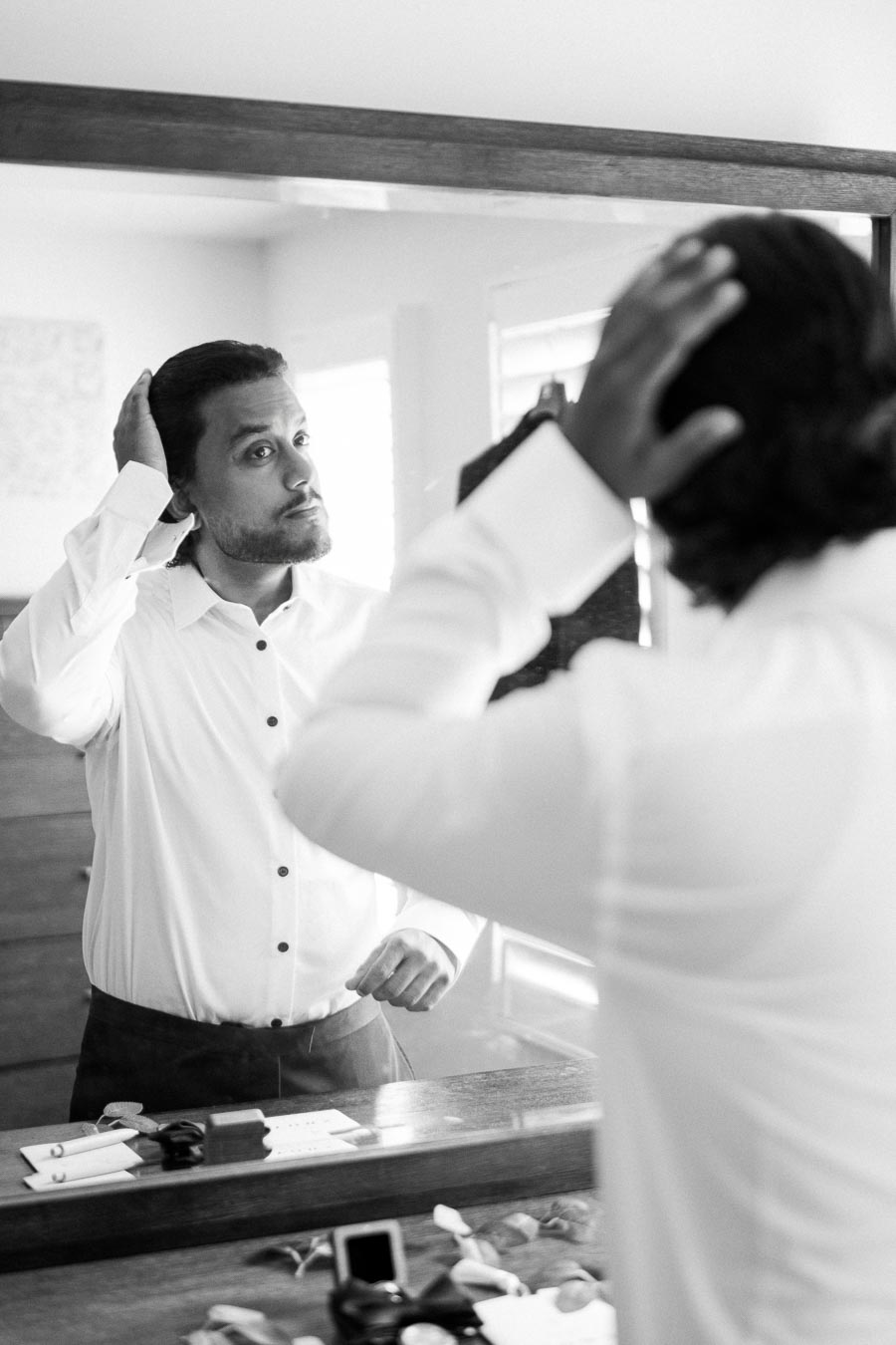 Black and white photo of a man adjusting his hair in front of a mirror, wearing a white dress shirt, preparing for a formal occasion or event.