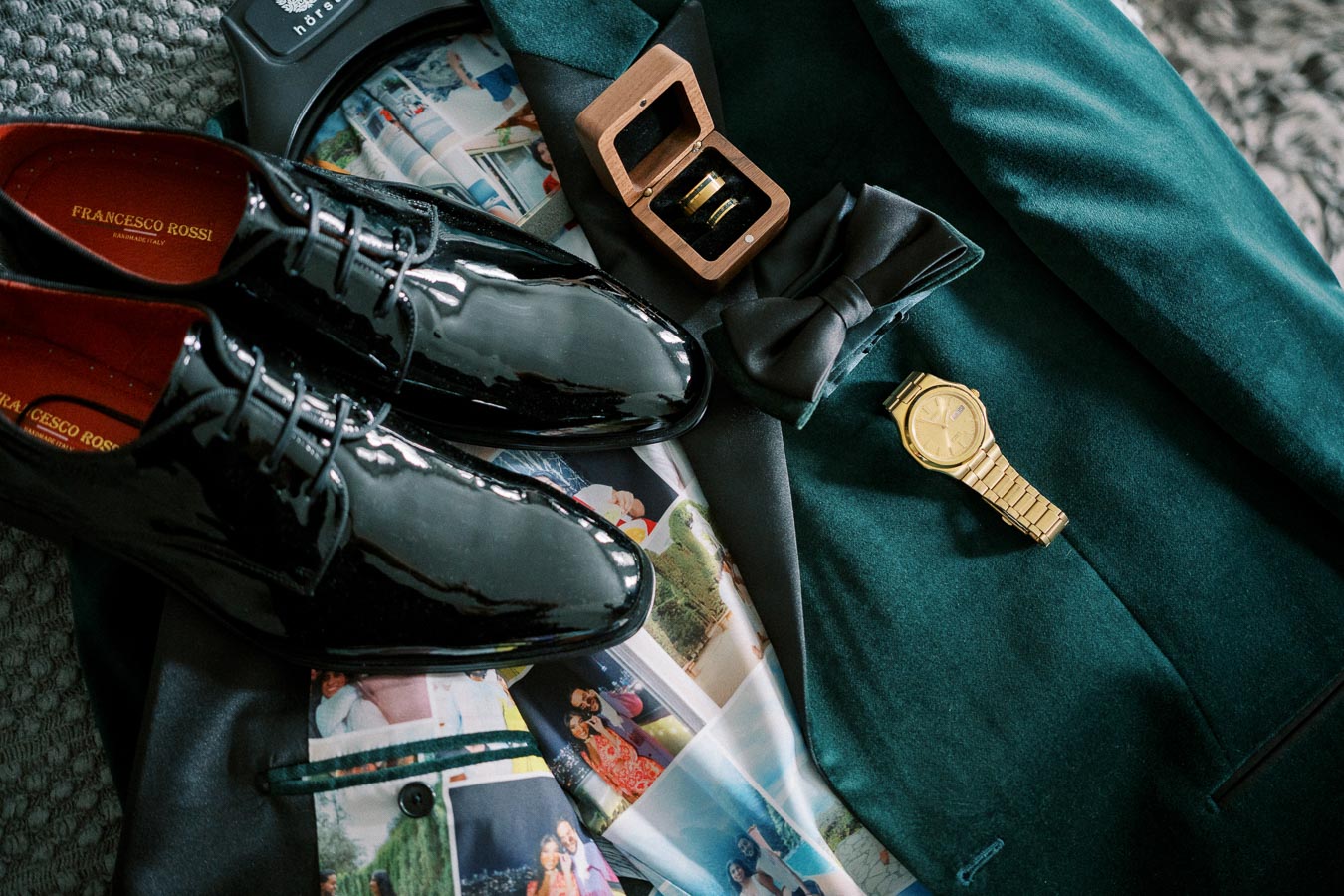 Elegant wedding attire layout featuring a dark green suit, black patent leather shoes, a gold wristwatch, and a bow tie, complemented by a ring inside a wooden box, ideal for a stylish groom's ensemble.