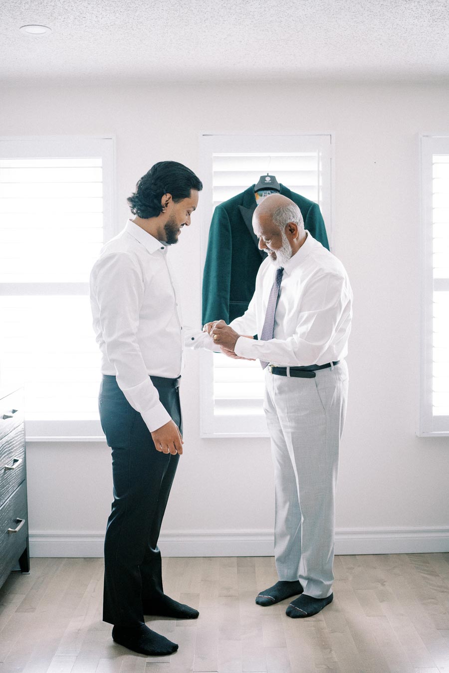 Two men in a bright room getting ready, one helping the other with his cufflinks, green jacket hanging in the background.