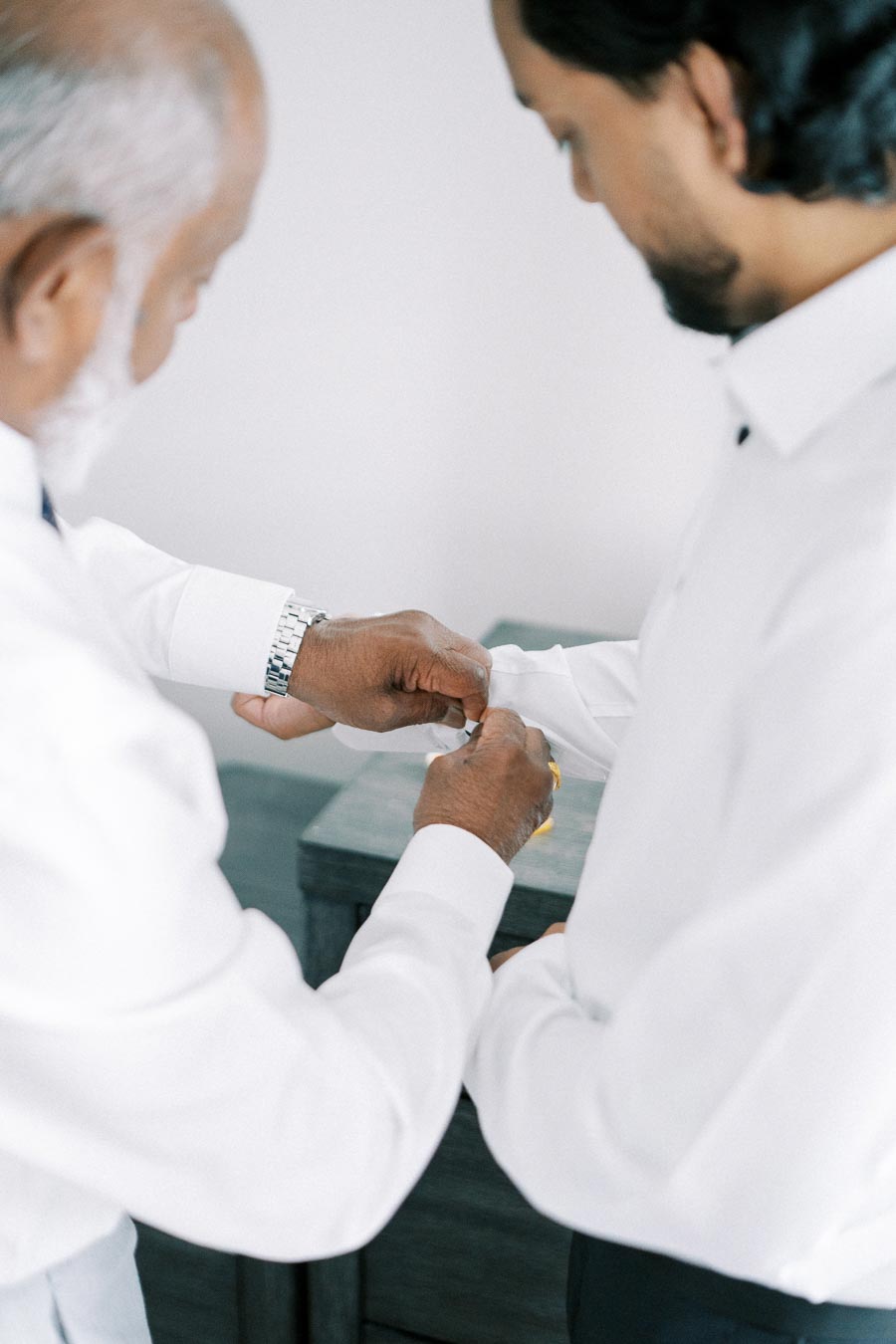 Two men in white dress shirts helping each other adjust cufflinks, focusing on the hands and attire, symbolizing cooperation and preparation.