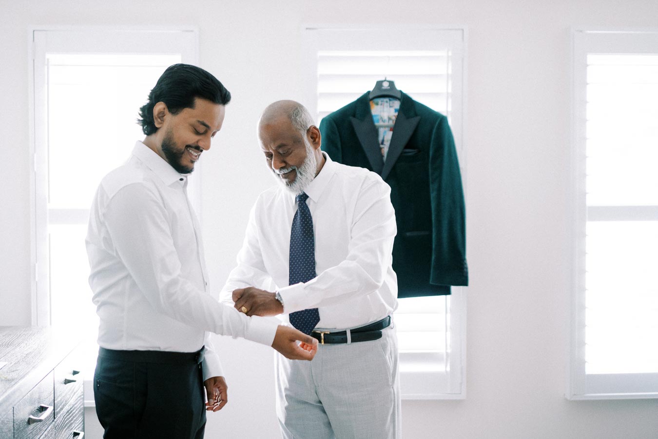 A younger and older man dressed in formal white shirts and ties, smiling and adjusting cufflinks in a bright room with a suit jacket hanging in the background.