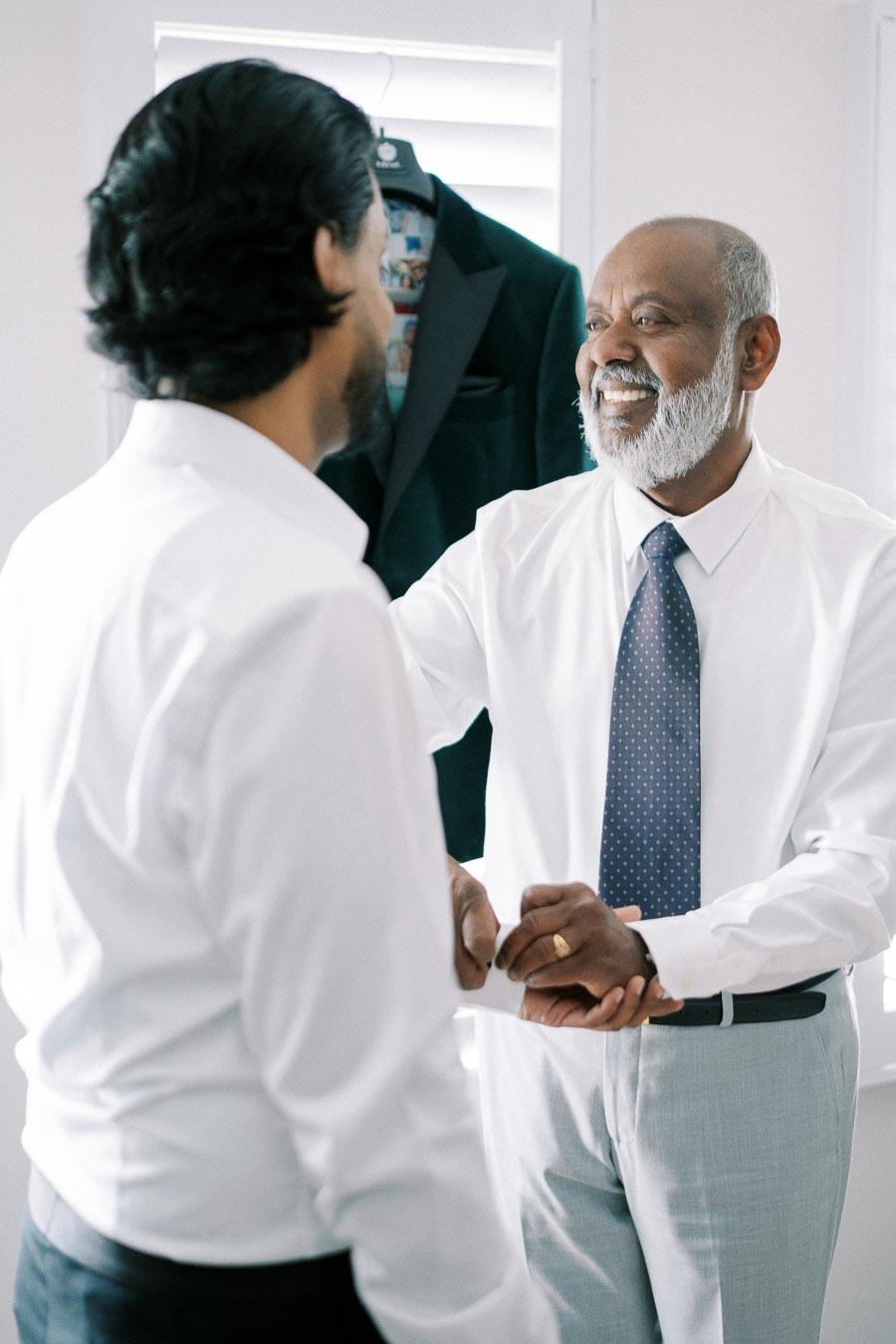Two men in formal attire, with one smiling and adjusting the other's sleeves, in a brightly lit room with a suit hanging in the background.