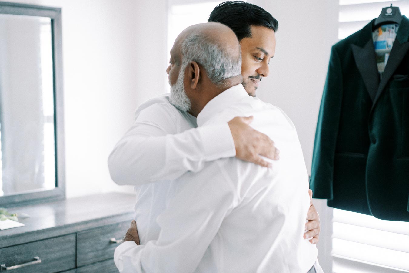 Two men in white shirts embrace in a heartfelt hug indoors, with a suit jacket hanging nearby, conveying a moment of connection and warmth.