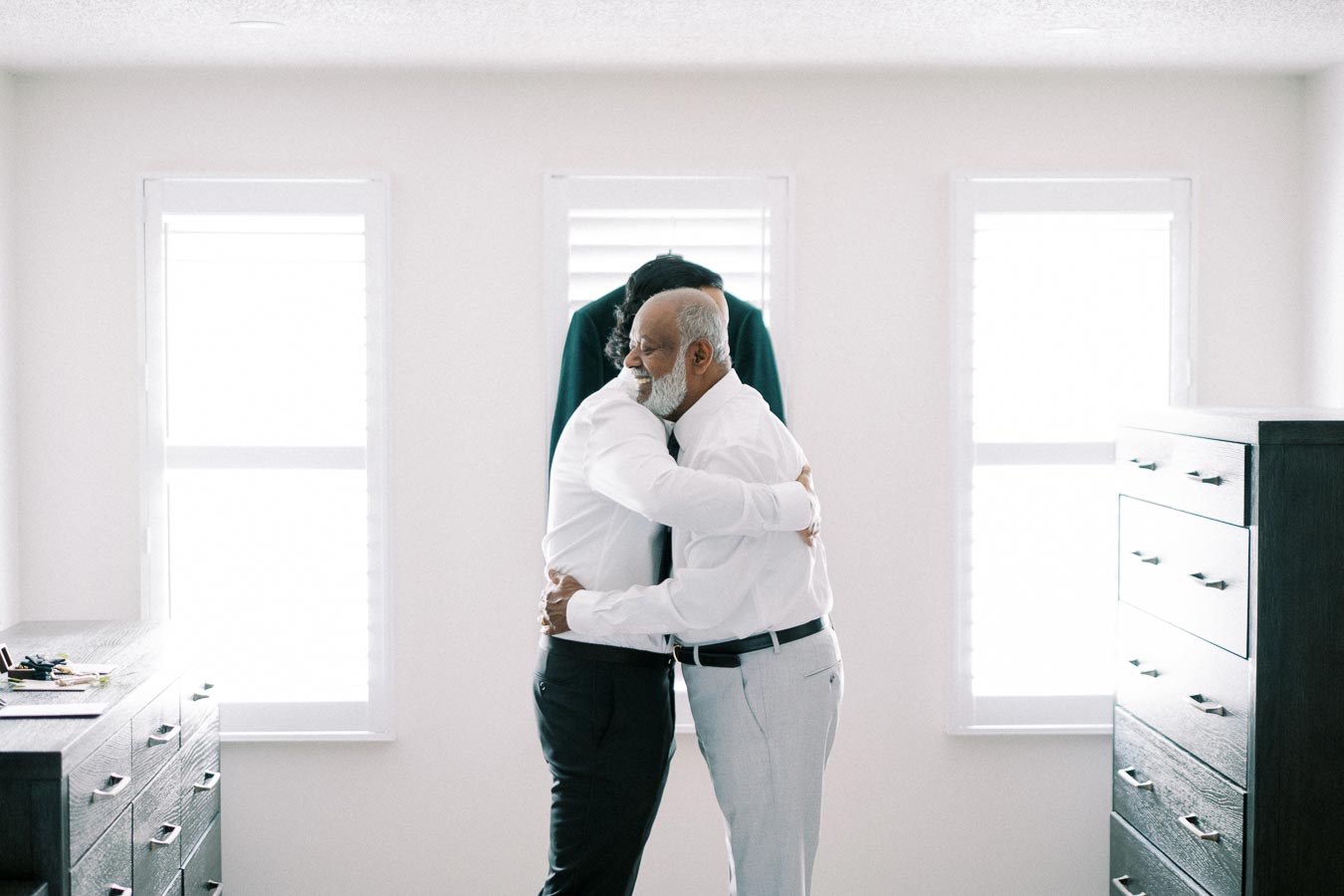 Two men hugging in a brightly lit room with dressers in the background, creating a warm and emotional moment.