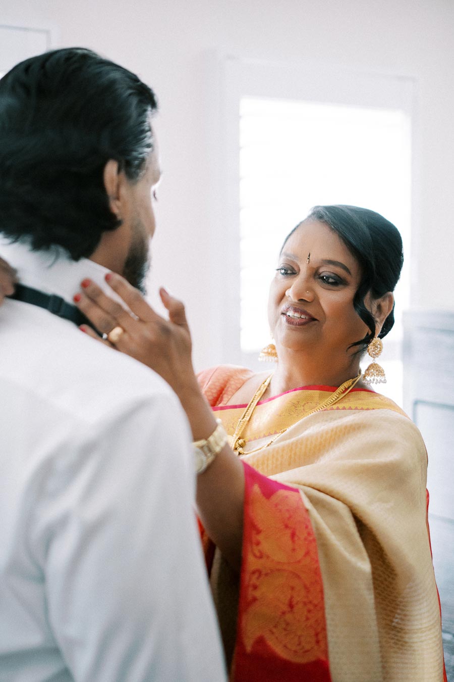 Mother adjusting son's bow tie on wedding day, both smiling warmly, she is wearing a golden sari.