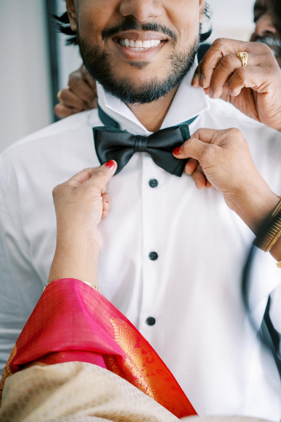 A groom in a white shirt gets his black bow tie adjusted by helping hands, highlighting the preparations for a traditional wedding ceremony.