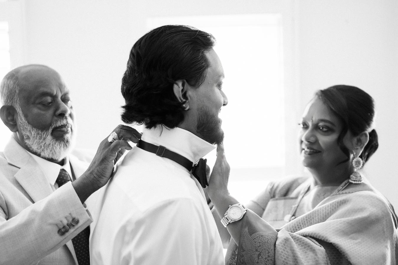 A black and white photo of a groom preparing for his wedding with the support of two elder relatives, adjusting his collar and smiling warmly in a sunlit room.