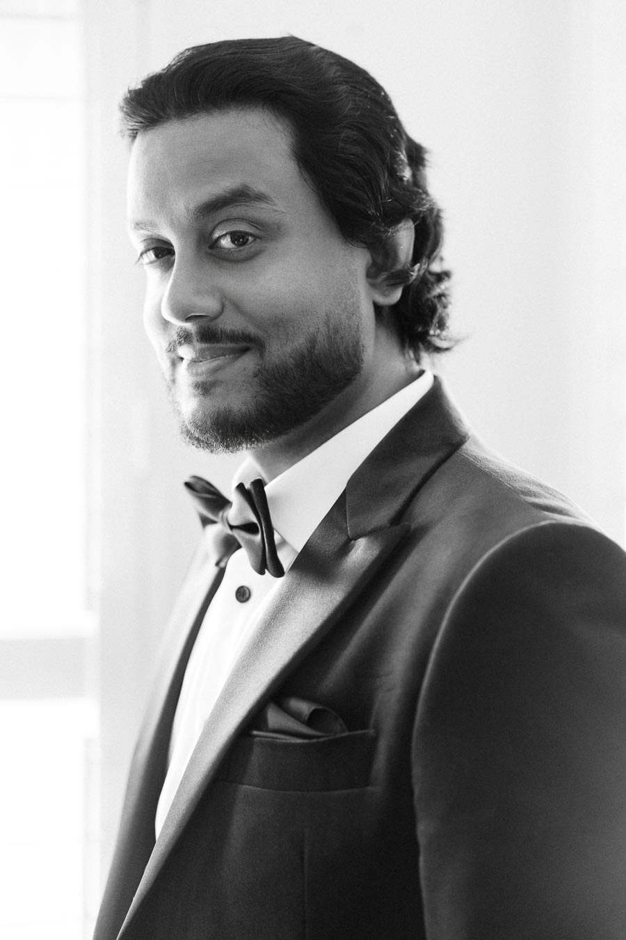 A man in a tuxedo smiles confidently, dressed formally with a bow tie, in a black and white portrait.
