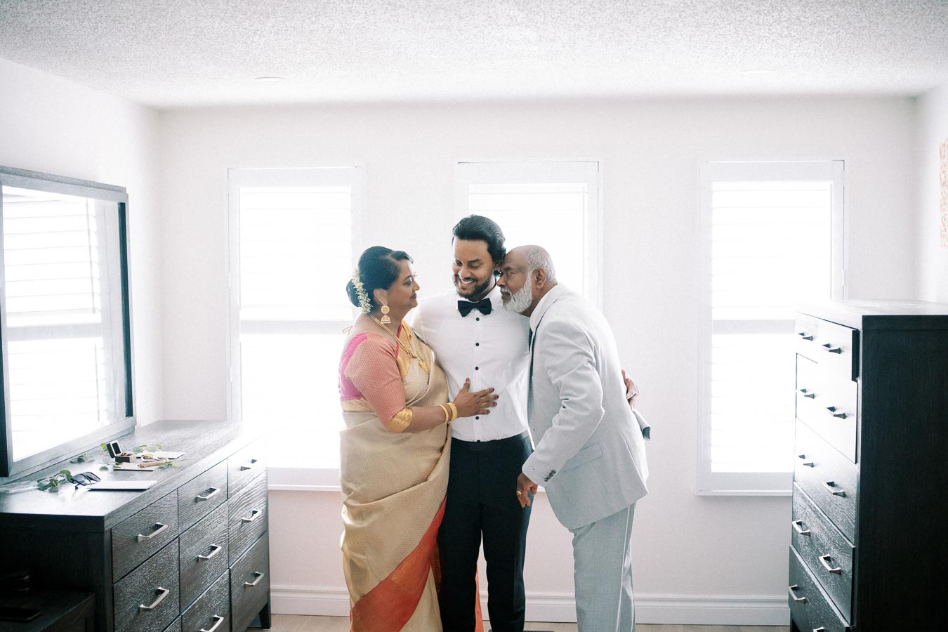 A groom in a white shirt embraces his parents, celebrating together in a warmly lit room with wooden furniture, highlighting a loving family moment.