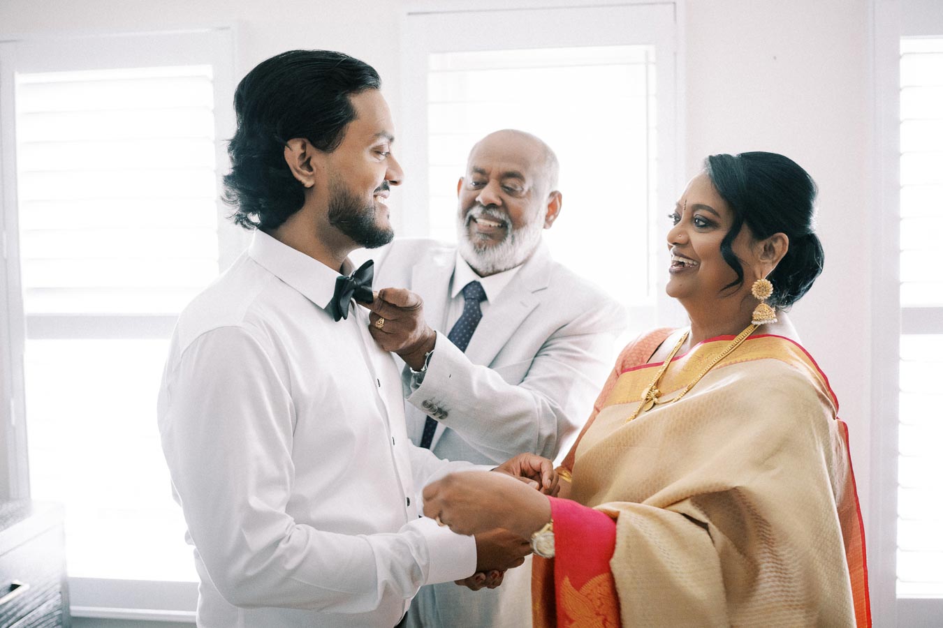 A joyful family moment as a man, likely the groom, gets ready for his wedding day with the assistance of his smiling parents in a sunlit room.