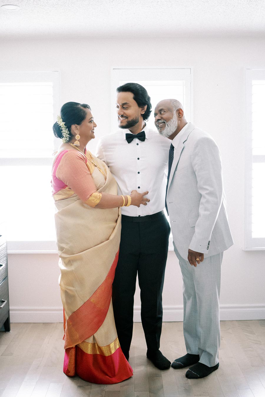 A groom in a tuxedo shares a joyful moment with his parents, who are wearing traditional attire, in a bright room with natural light pouring in through the windows.