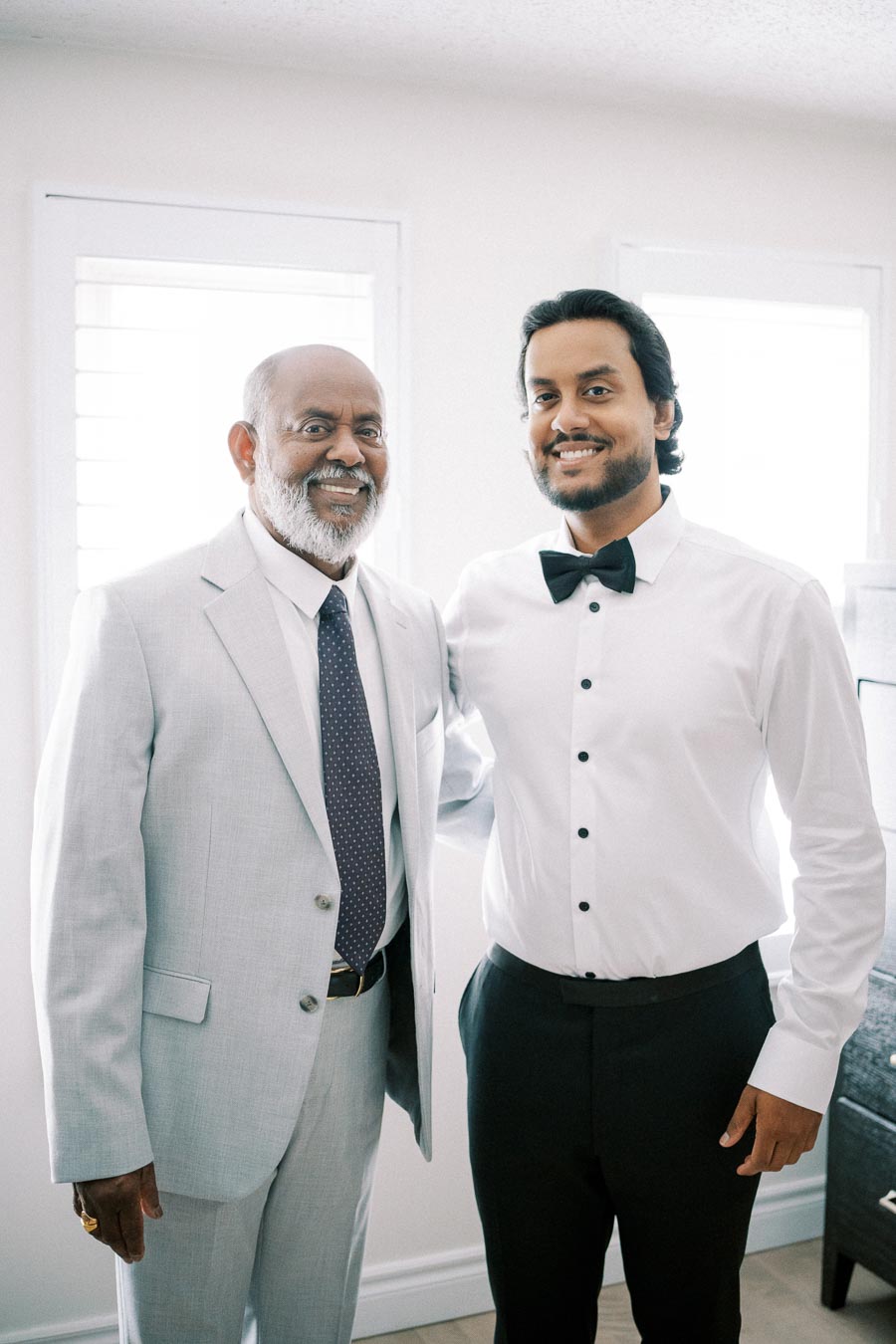 Two men dressed formally in a well-lit room, one in a light gray suit and the other in a white shirt with a black bow tie, smiling at the camera.