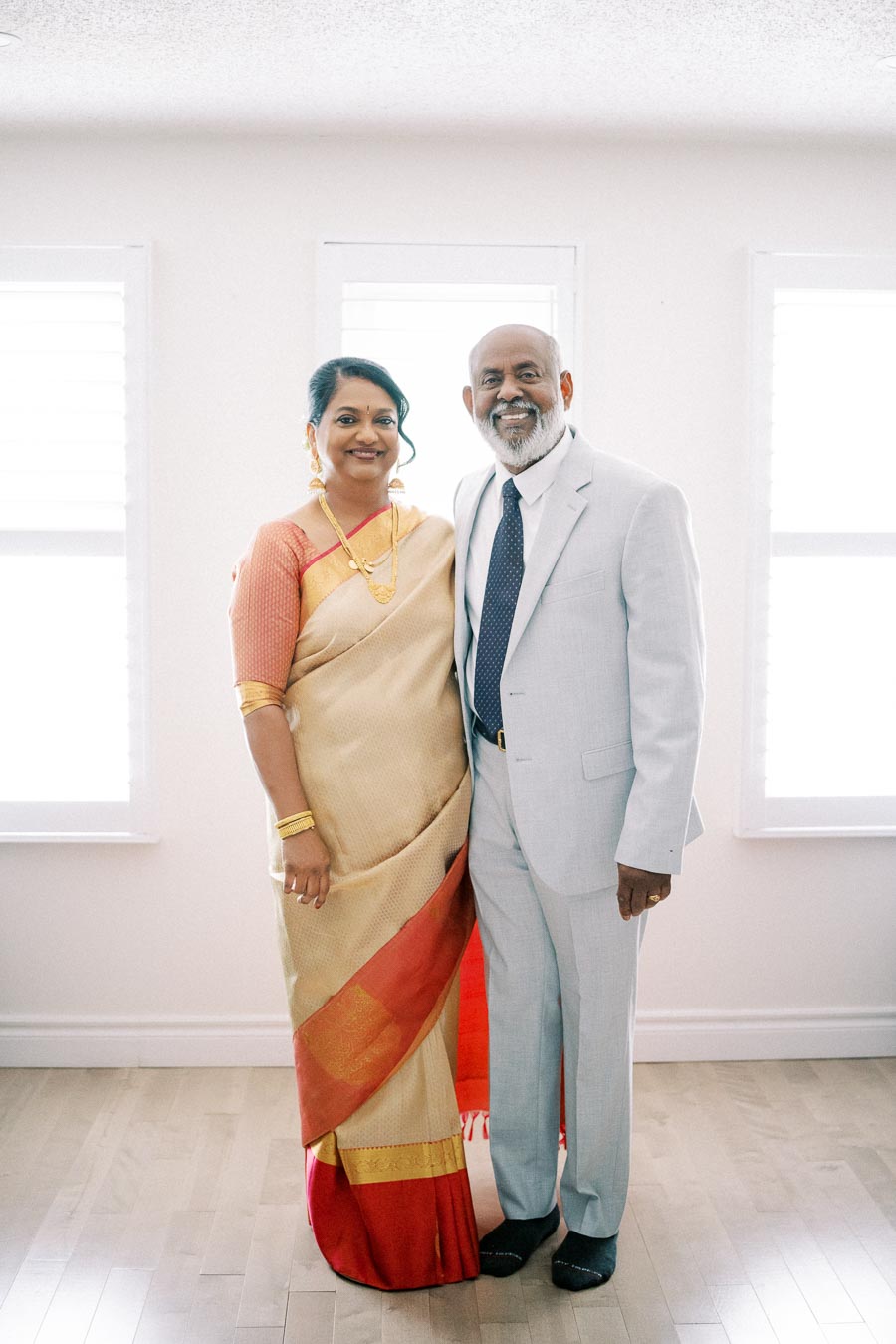 Elderly couple dressed in formal attire, with the woman wearing a traditional sari and the man in a light-colored suit, standing together smiling in a well-lit room.
