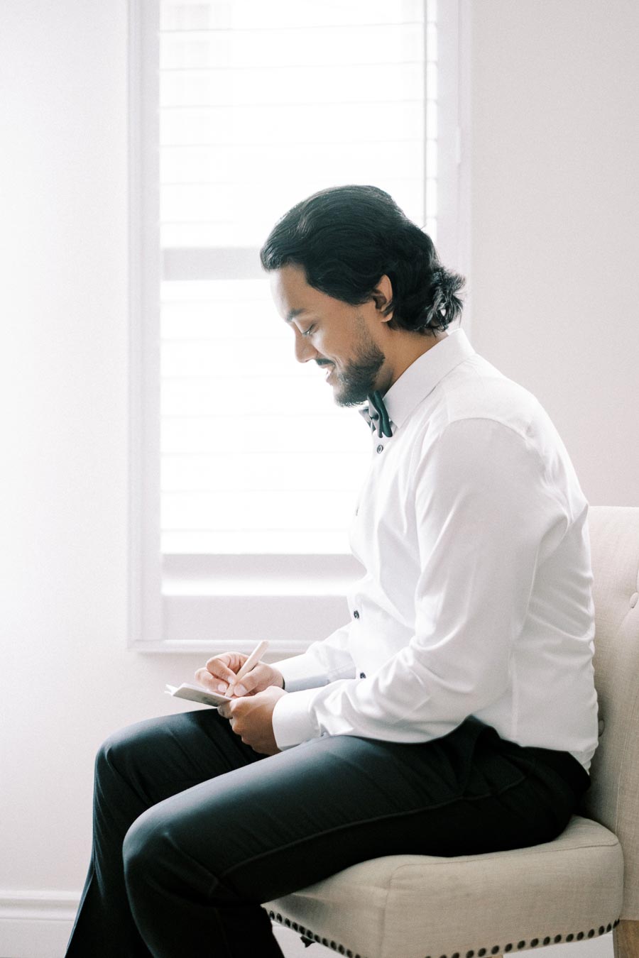 Man in formal attire writing in a notebook while seated near a well-lit window.