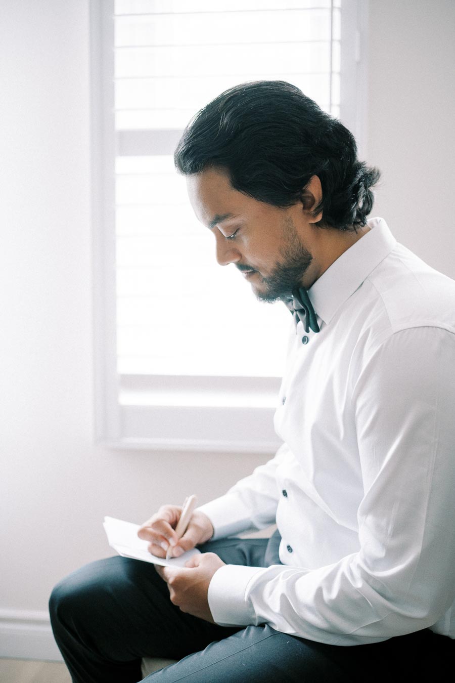 Young man in a formal white shirt writing a letter while seated by a bright window.