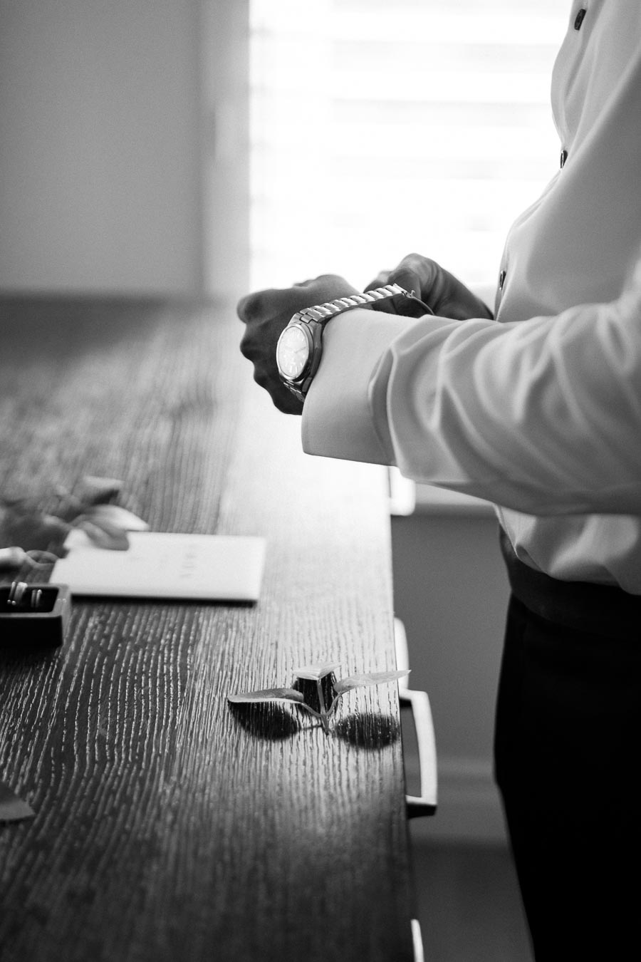Black and white image of a man in a dress shirt adjusting his wristwatch at a wooden table, with a ring box and papers nearby, symbolizing preparation and elegance.