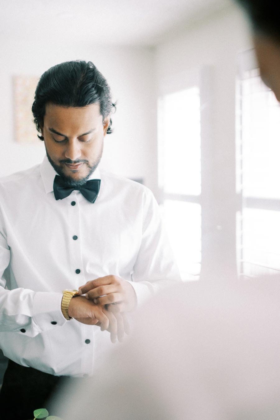 A man in a white dress shirt and black bow tie adjusts his gold wristwatch while standing in front of a bright window, preparing for a formal event.