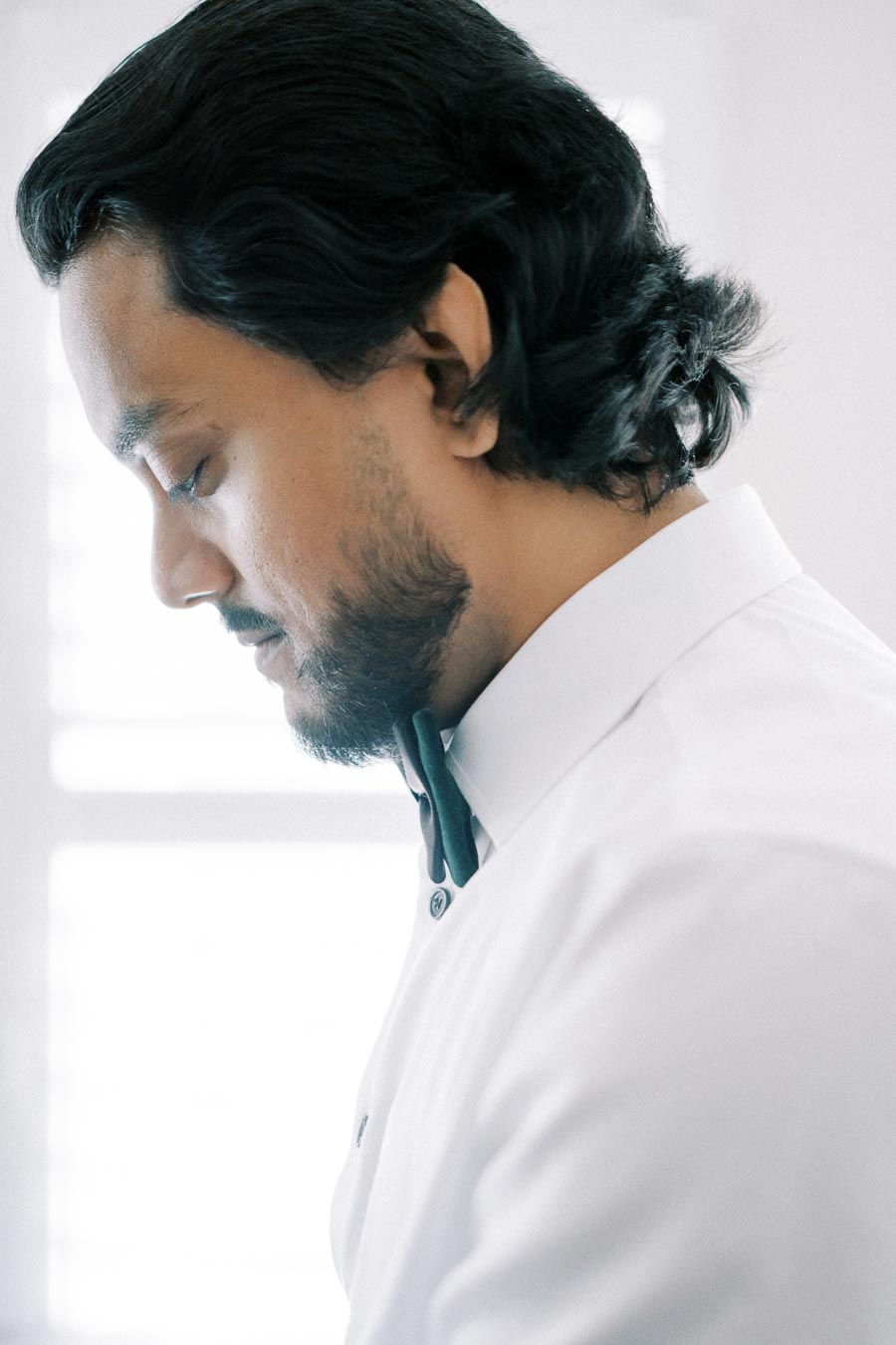 Man in a white dress shirt and black bow tie looking downward in a softly lit room.