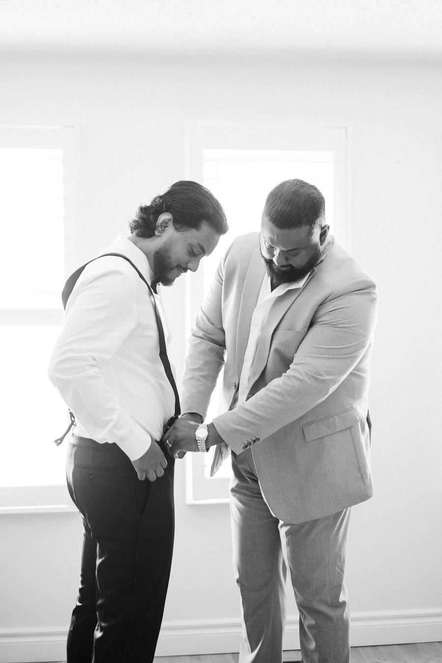 Two men in formal attire getting ready, one adjusting the other's suspenders in a brightly lit room, preparing for a formal event.