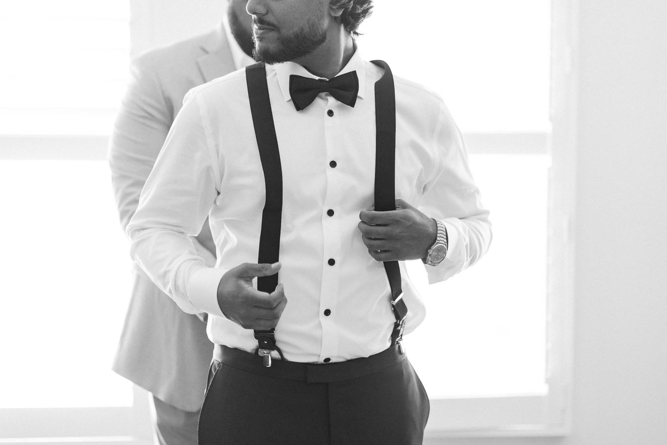 A groom adjusting his suspenders while wearing a white dress shirt, black bow tie, and a watch, preparing for his wedding day in a bright room.